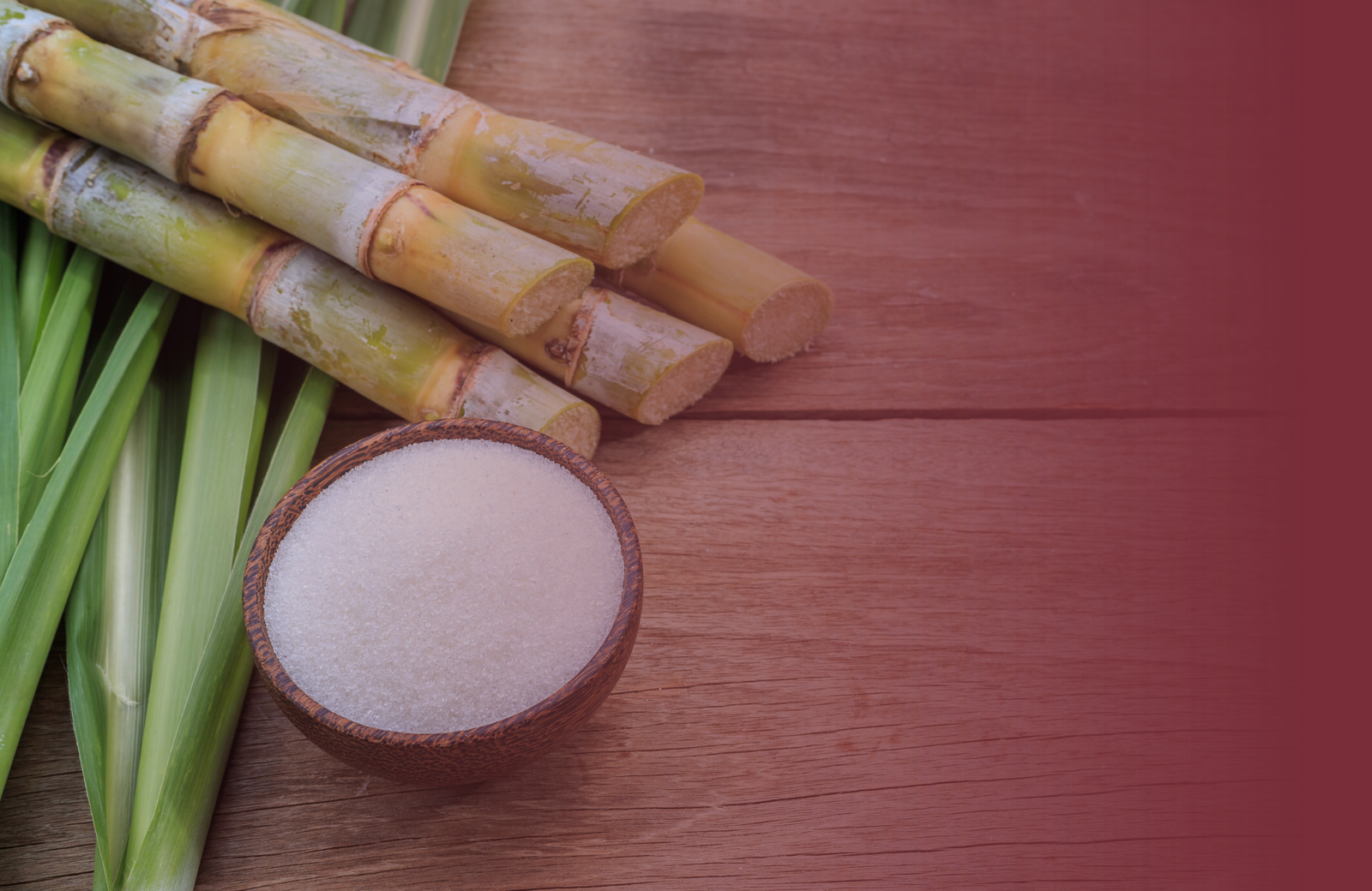 Sugarcane stalks and a bowl of sugar on a maroon wooden surface
